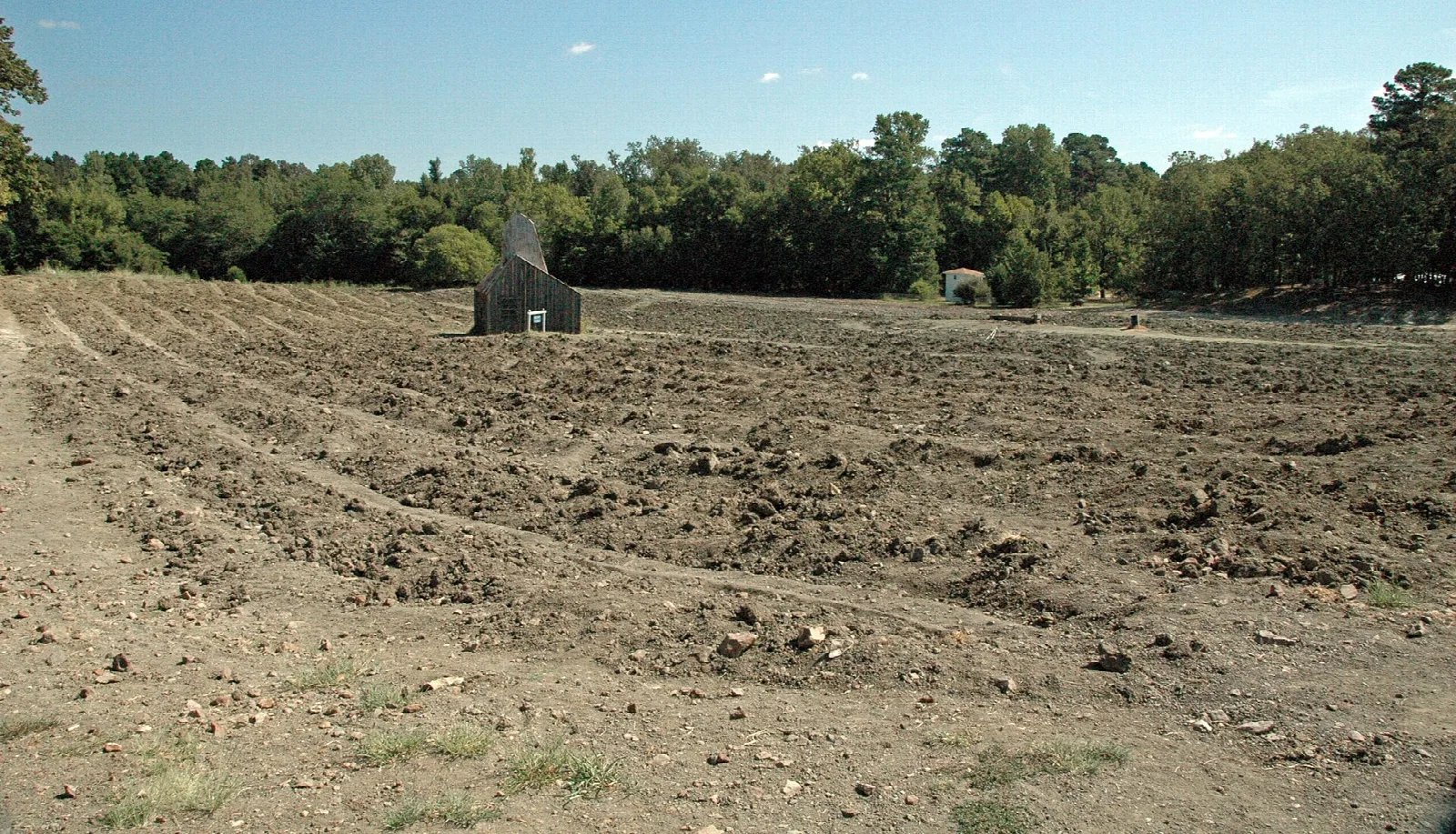 Wide plowed dirt field with a small shed in the distance and forest beyond