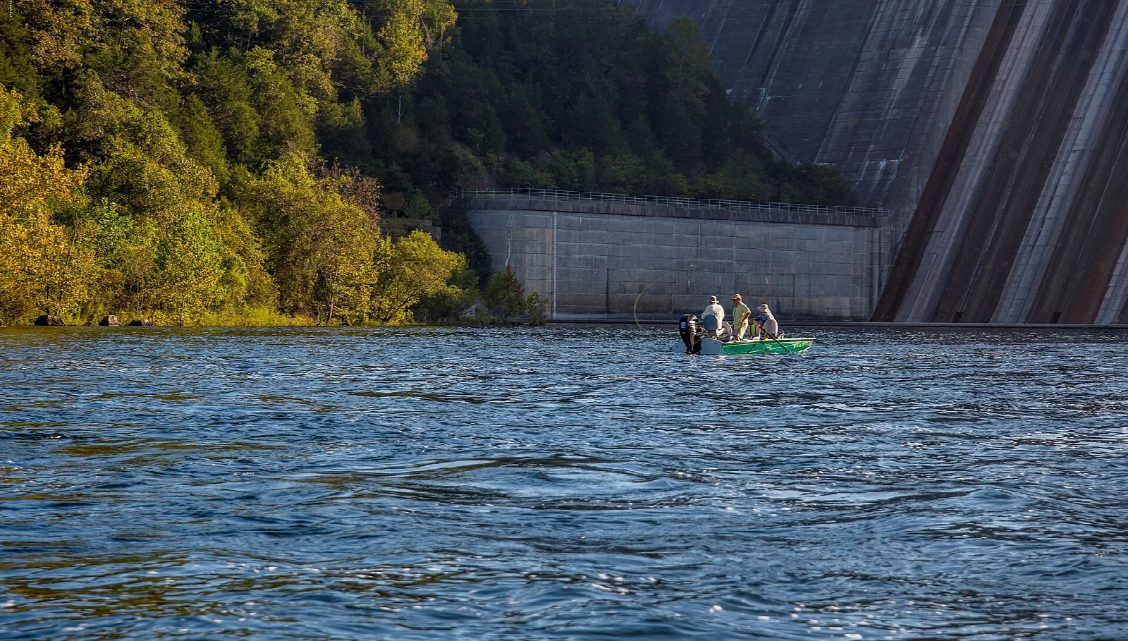 Small boat with anglers on the river below a tall concrete dam wall