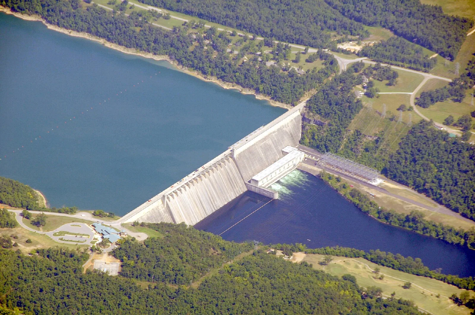 Aerial view of a dam between a blue lake and a river valley with forest and fields