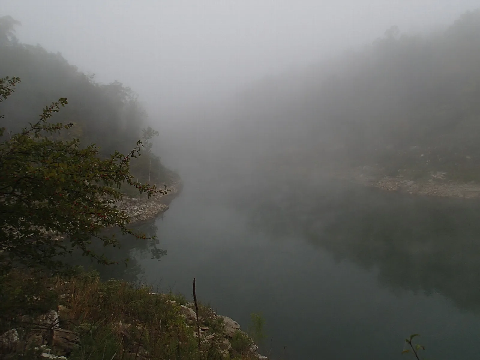 Fog over a quiet lake edge with rocky shore and wooded hills
