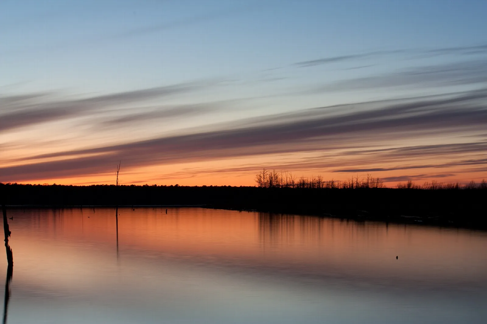 Calm lake at sunset with orange sky reflected in the water and a dark treeline