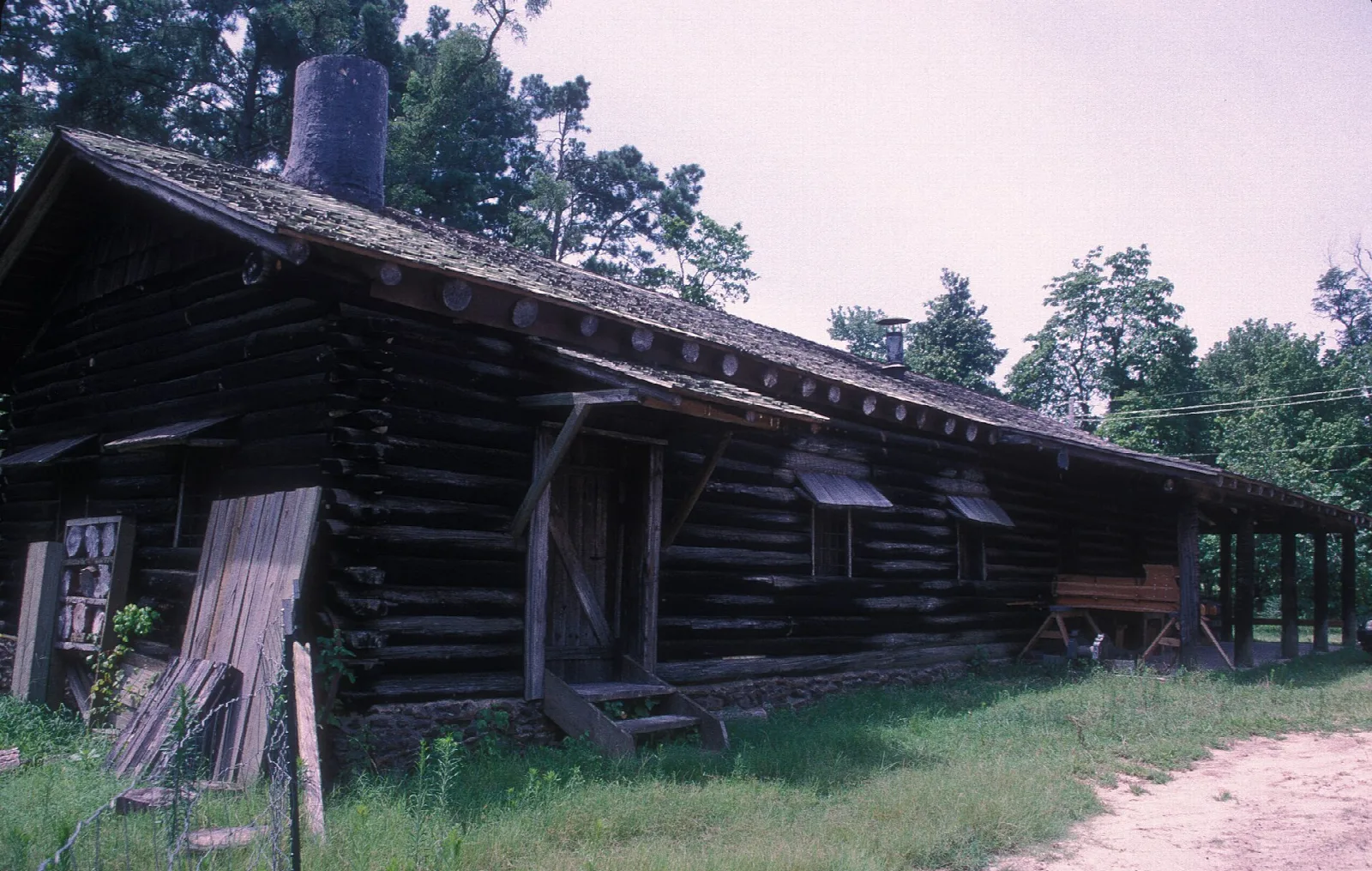 Weathered log building with a shingled roof and covered porch beside a dirt path