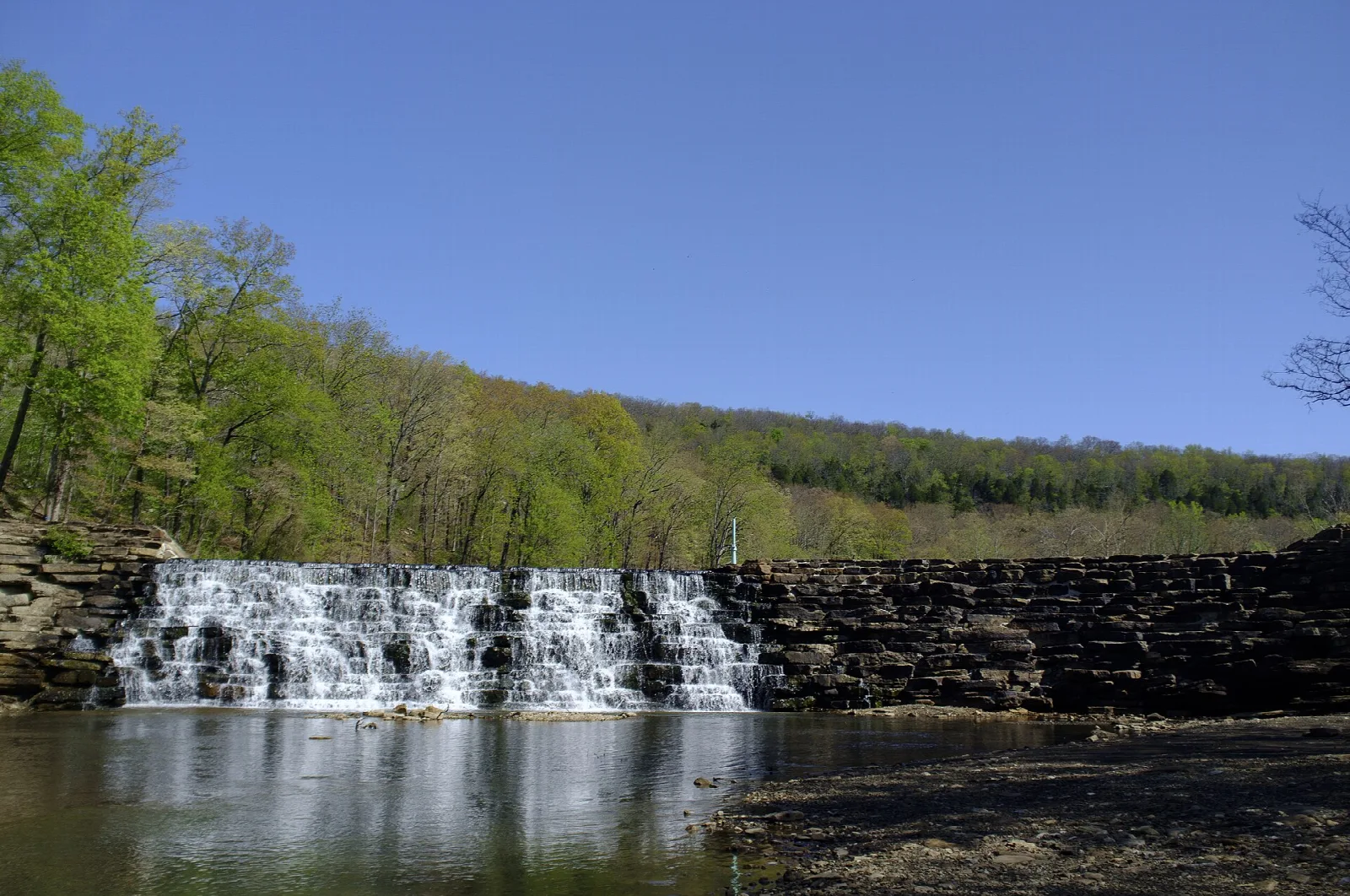 Low stone dam with water spilling over it into a pond beneath a blue sky and wooded hills
