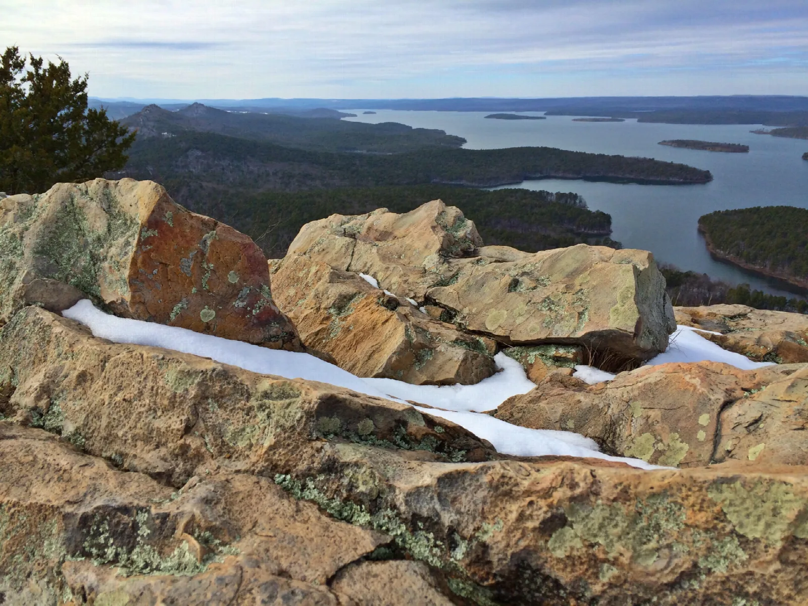 Rocks with patches of snow in the foreground and a lake among forested hills beyond