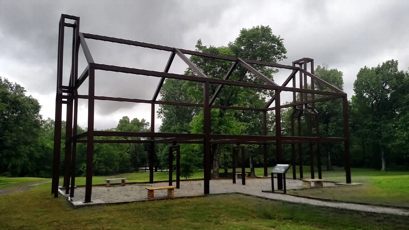 Dark steel frame outlining a building on a grassy lawn with benches and trees