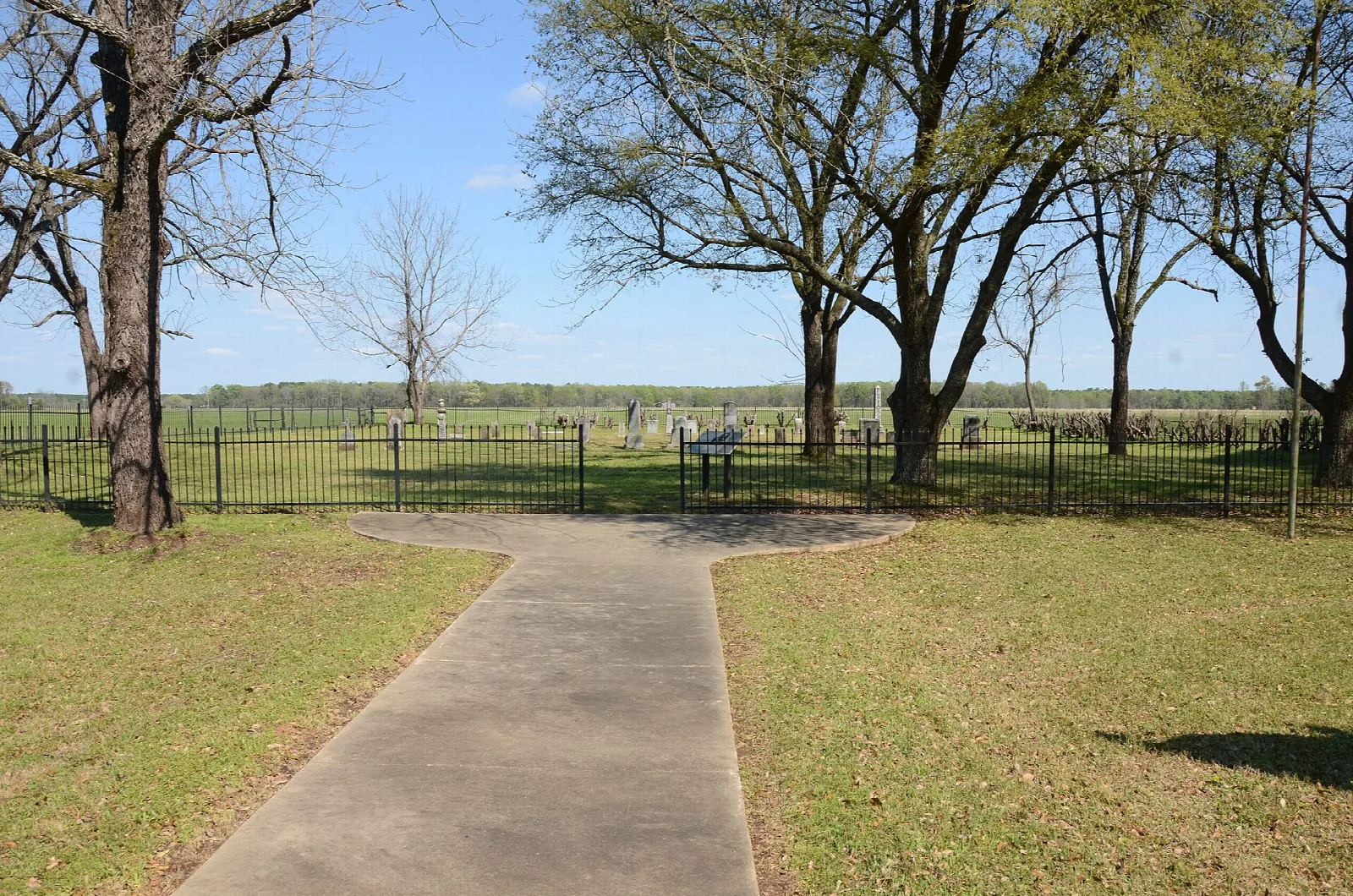 Paved path leading to a fenced cemetery in an open grassy field with trees