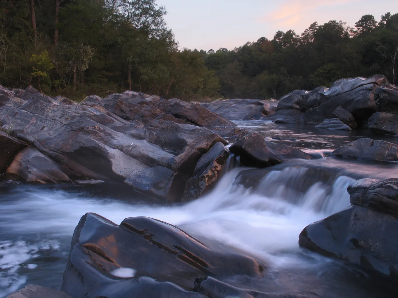 Water spilling over dark rocks at sunset in a wooded river