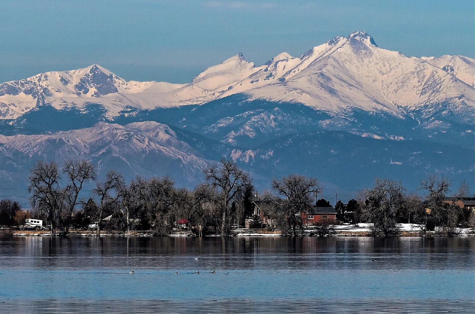 Snow-capped Rocky Mountain peaks across a blue lake with bare trees along the shore