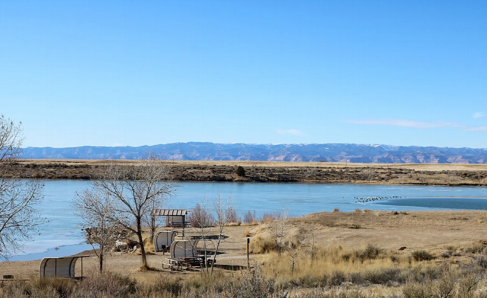 A lake edge with picnic shelters and far mesas under clear blue sky