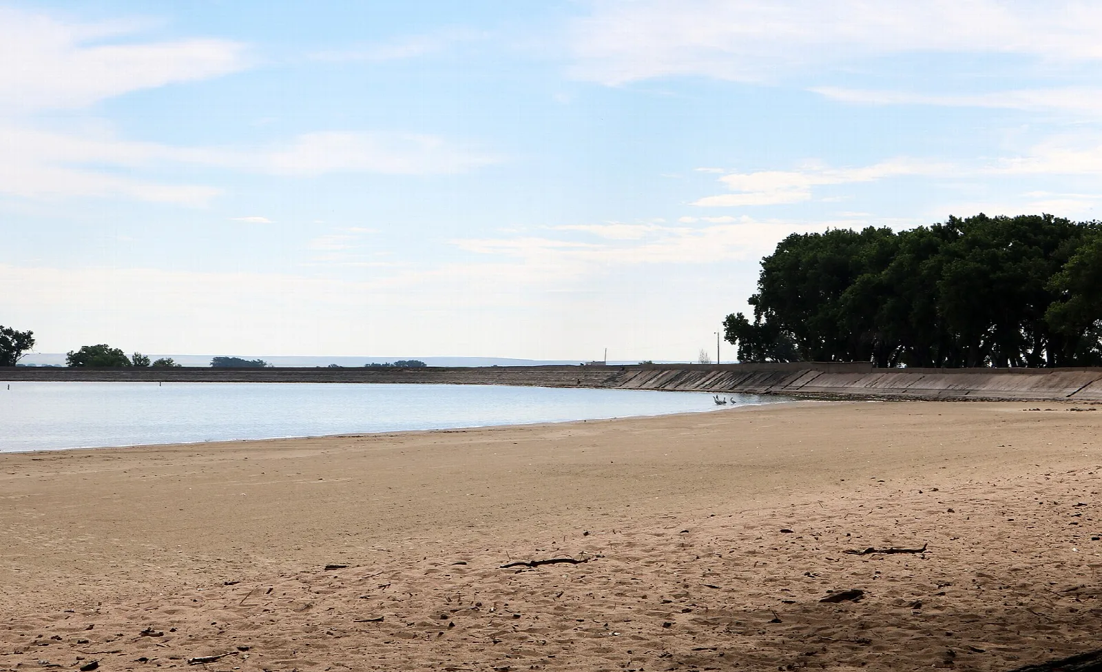 Bare sandy shore beside calm water, with a long low dam and trees in the distance