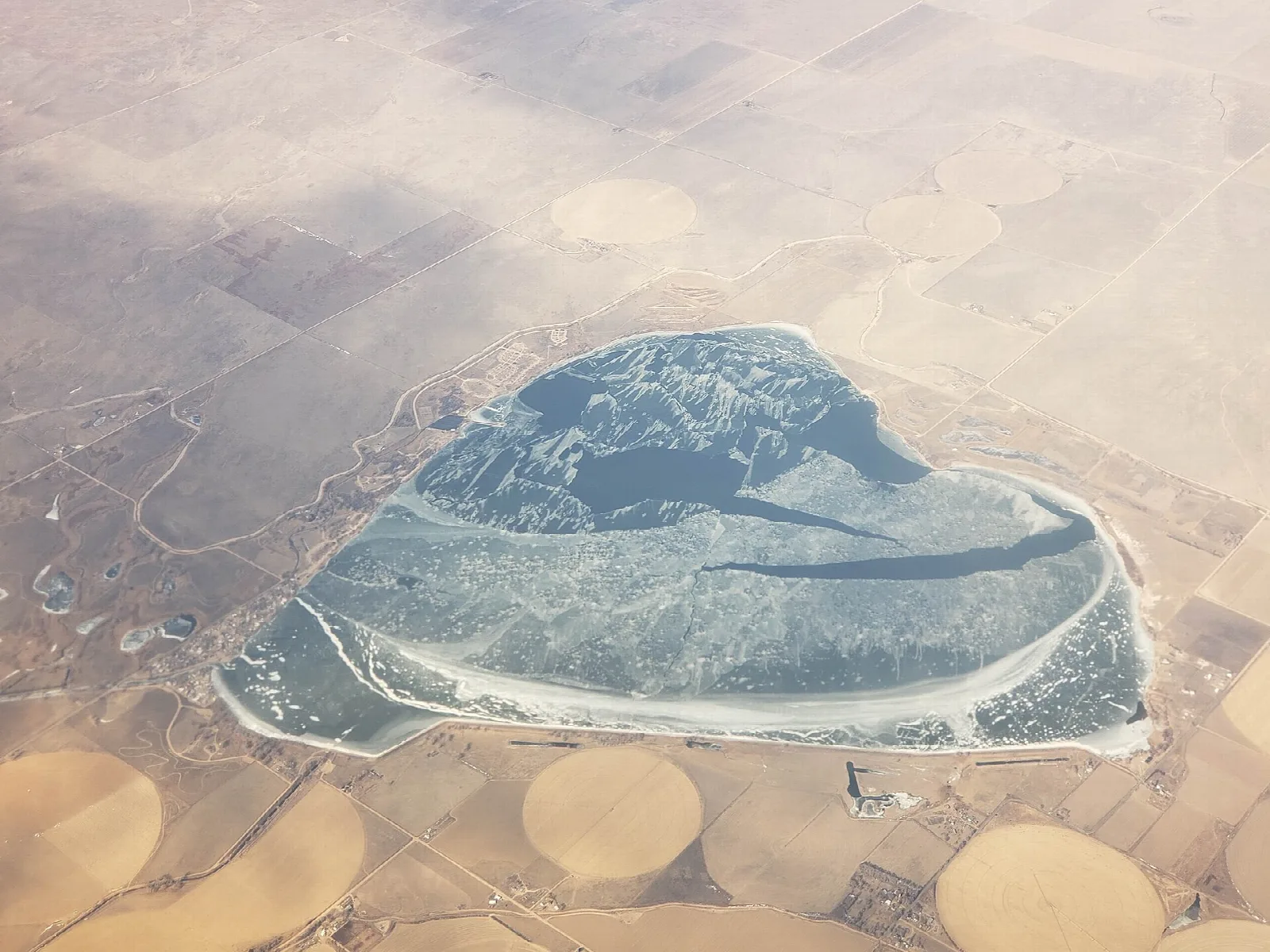 Aerial view of a partly iced reservoir surrounded by round farm fields