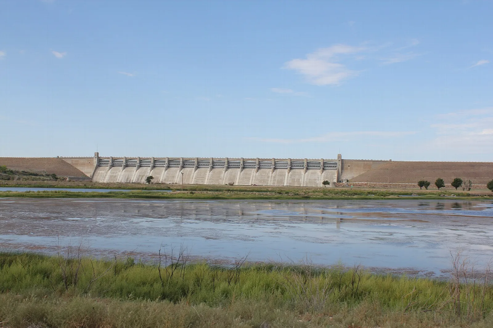 Long concrete dam beyond shallow water and green grass under blue sky