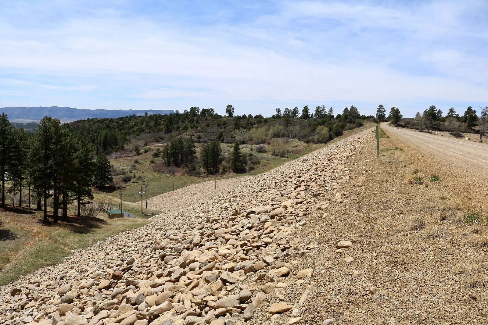 Gravel dam road beside a lake with pine forest and hills