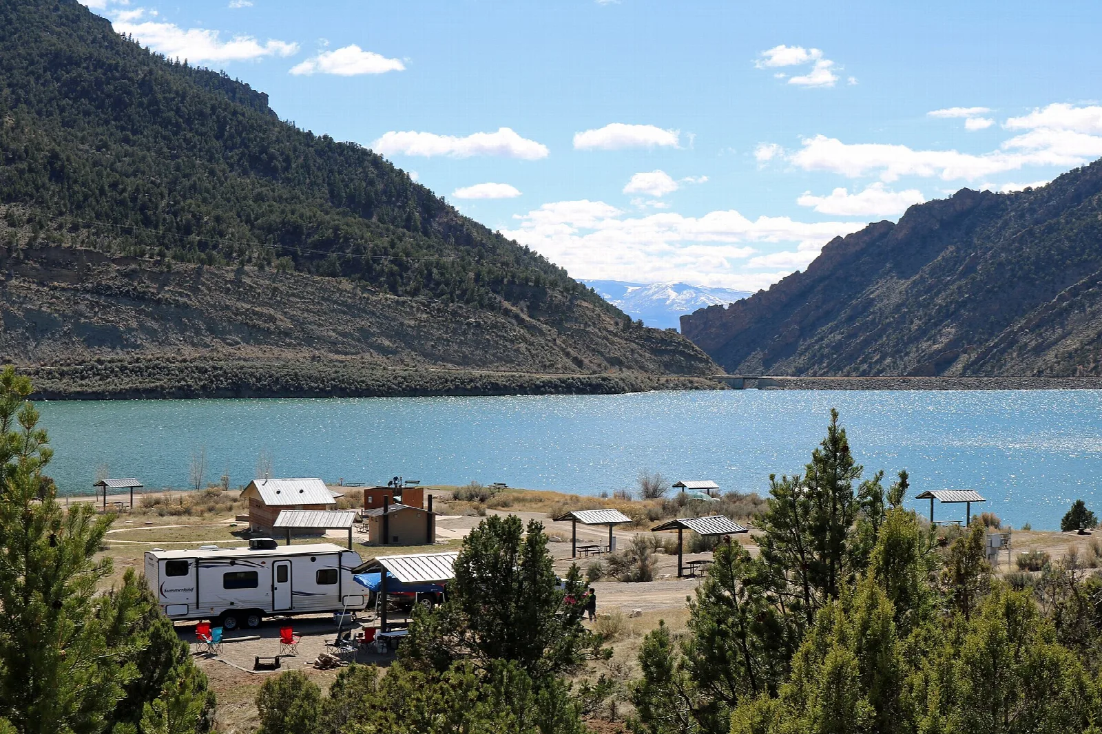 Turquoise reservoir below steep brushy hills, with a camper and picnic shelters in the foreground