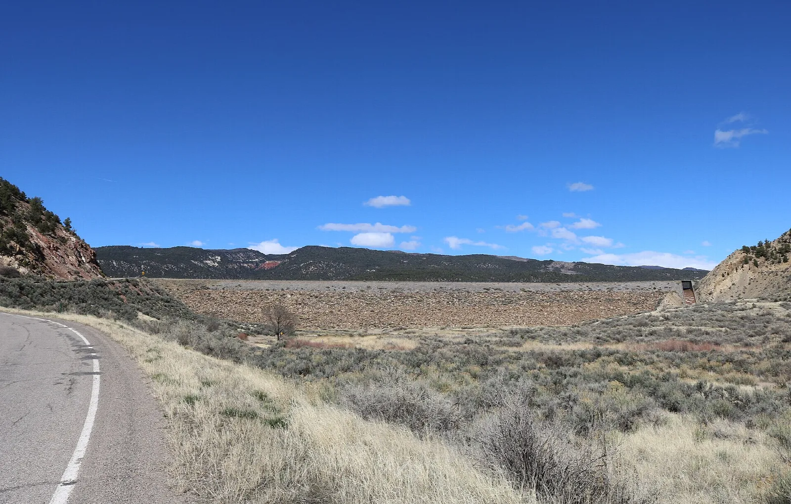 A paved road beside sagebrush leading toward a long earthen dam under a blue sky