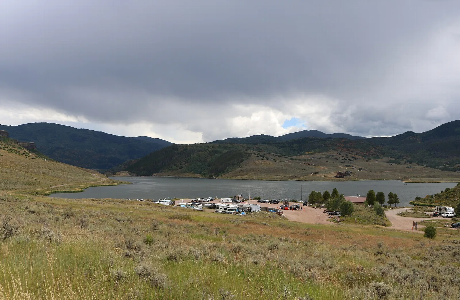 Lake under a stormy sky with a cluster of vehicles on the shore and hills beyond
