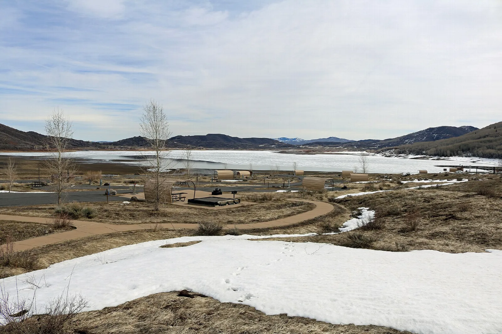 Partly frozen reservoir with a paved loop road and low hills beyond, patches of snow