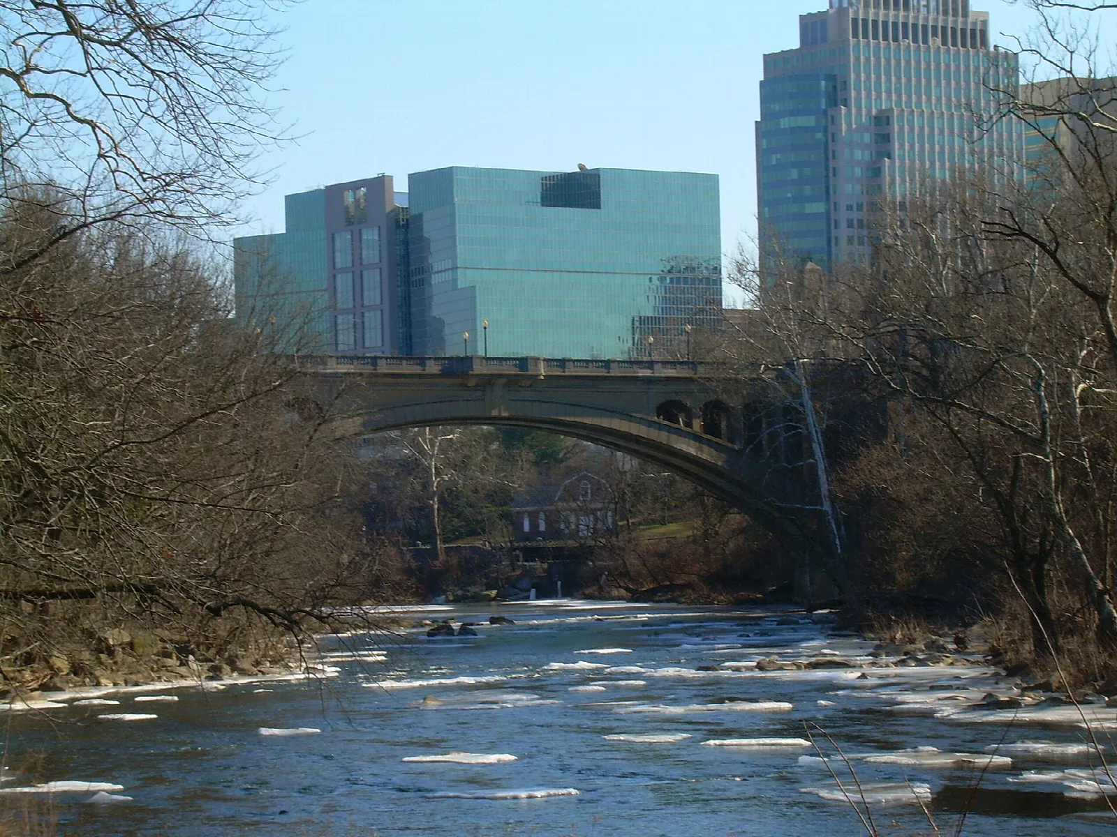 Creek with ice patches under a bridge, with glass office towers behind