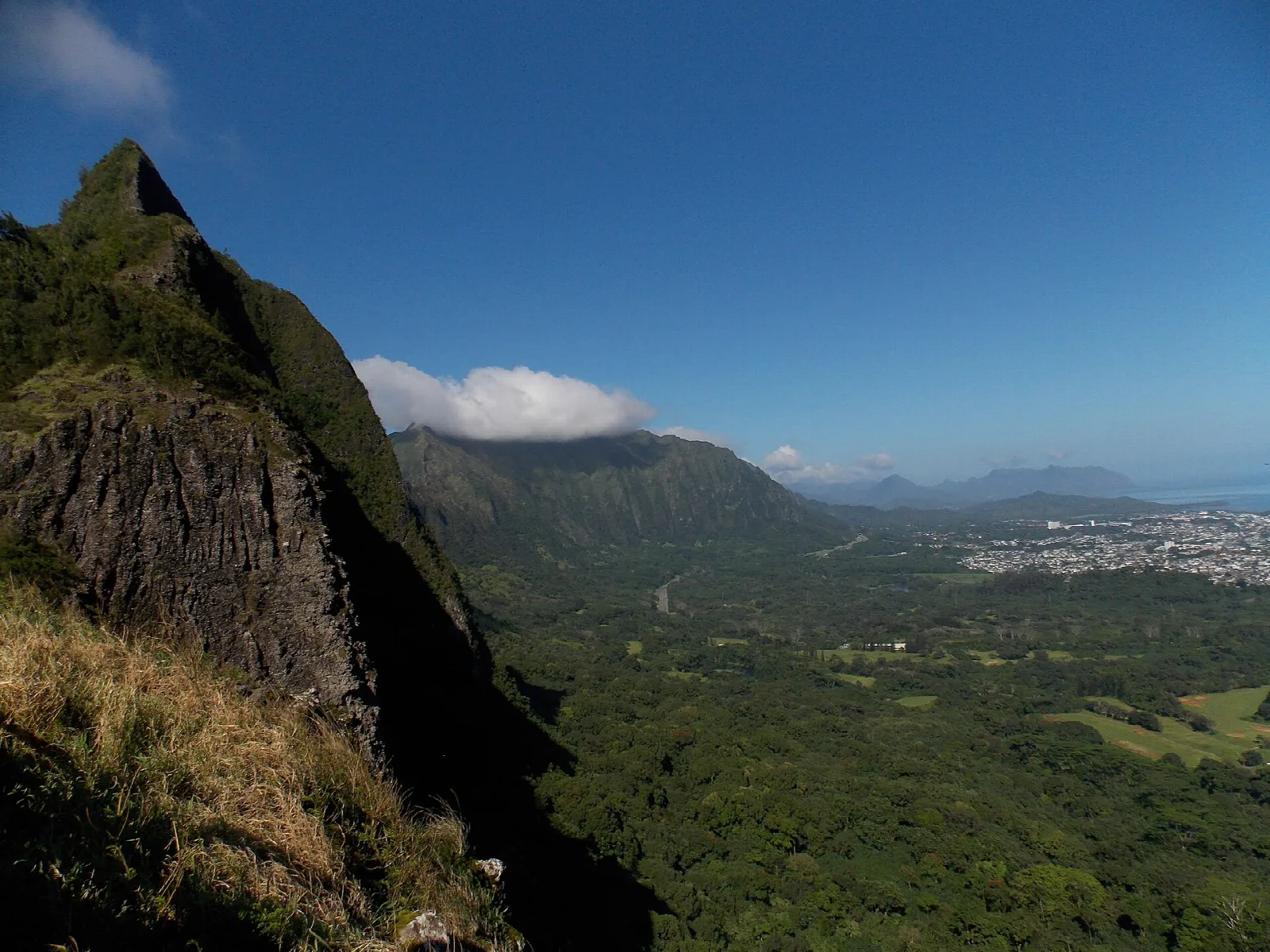 Sharp cliff peak at left above a green valley and far-off town and sea