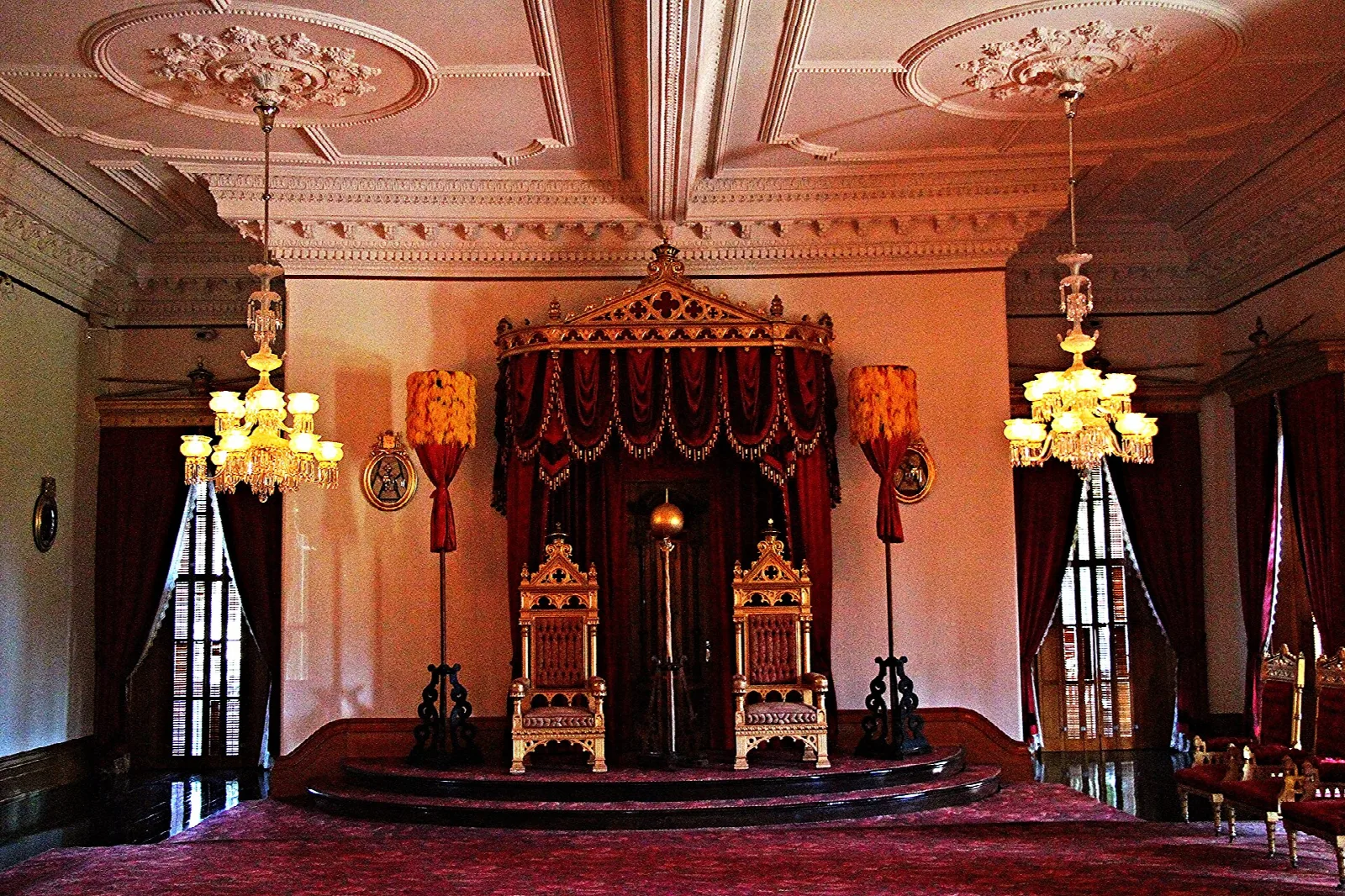 Throne room with two thrones under a red canopy and chandeliers