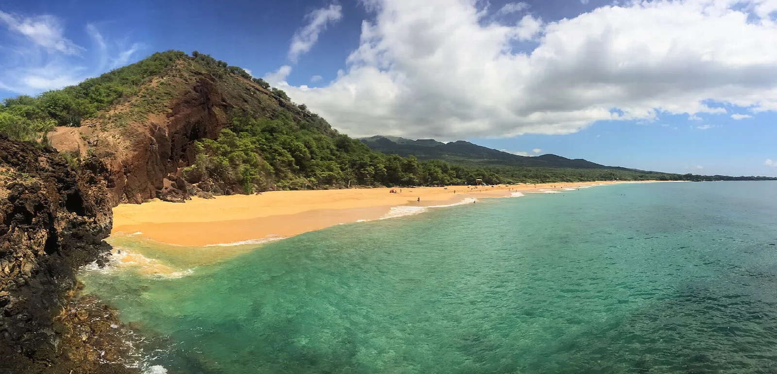 Golden beach curving beside clear green-blue water below a red cliff