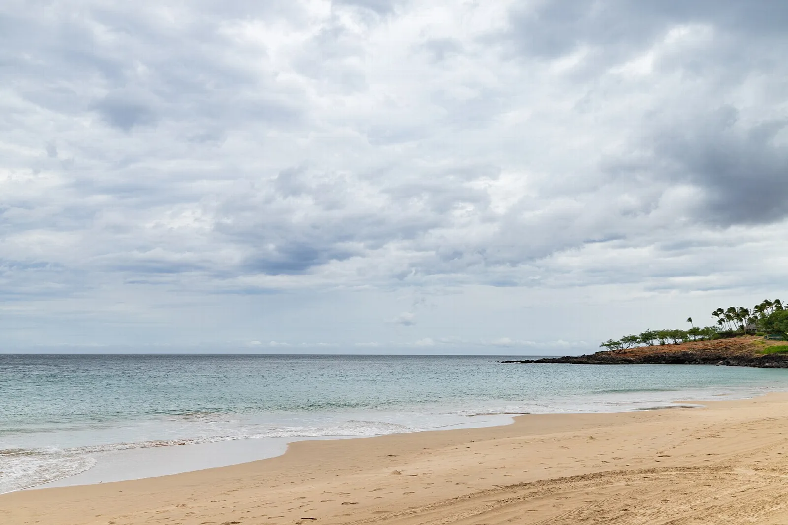 Pale sand and calm water below cloudy skies, with a wooded point at right