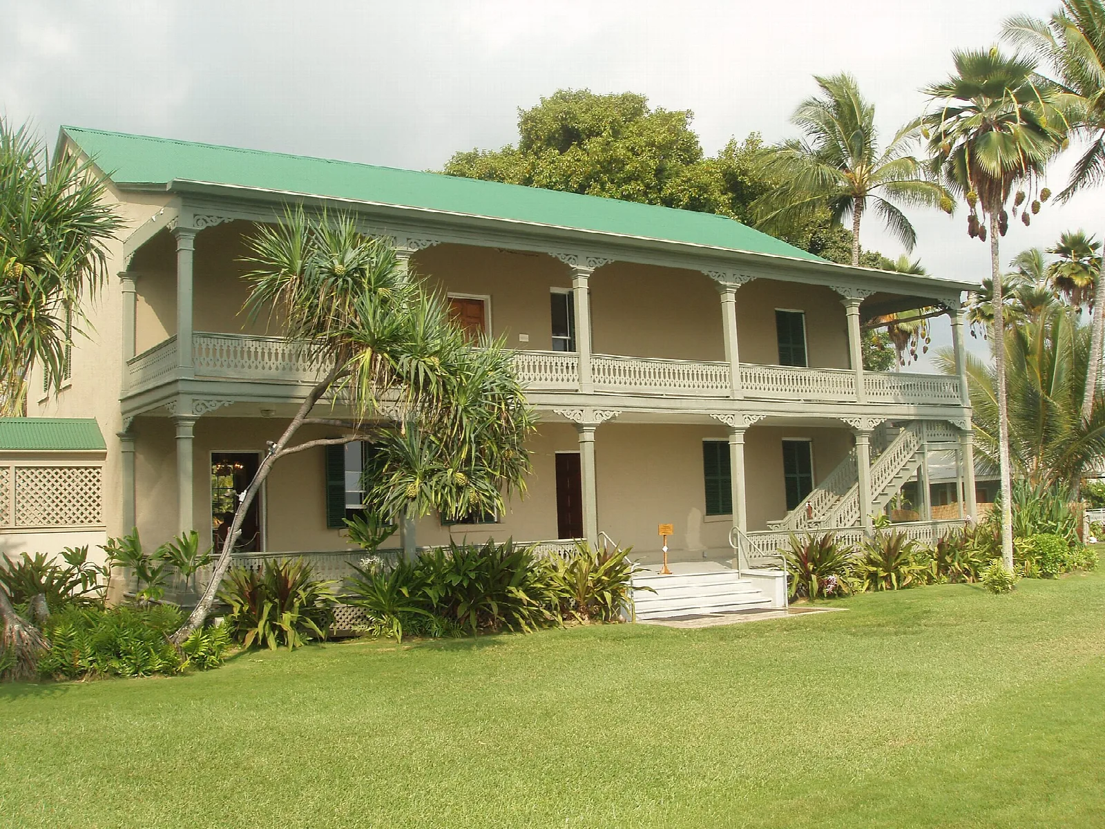 Two-story palace with a green roof and veranda beside a lawn and palms