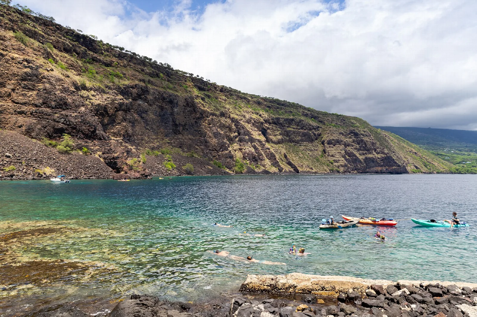 Snorkelers and kayakers in calm water below a steep volcanic cliff