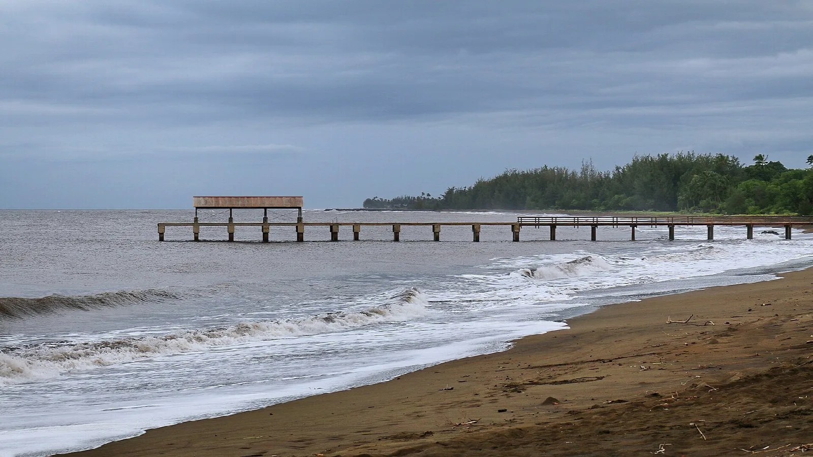 A pier reaching into the sea beside a dark sand beach