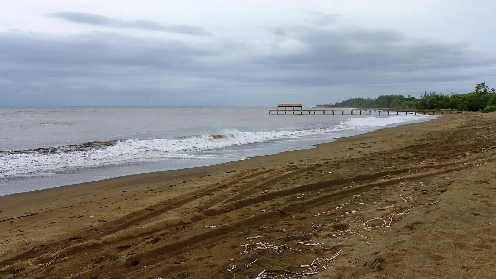 A dark sand beach with a pier in the distance under cloudy skies