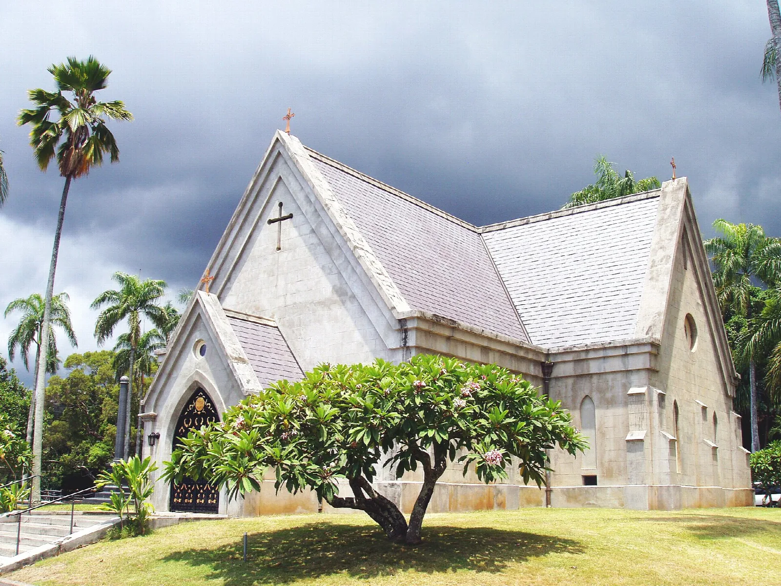 A gray stone chapel with a steep roof beside palms and a plumeria tree