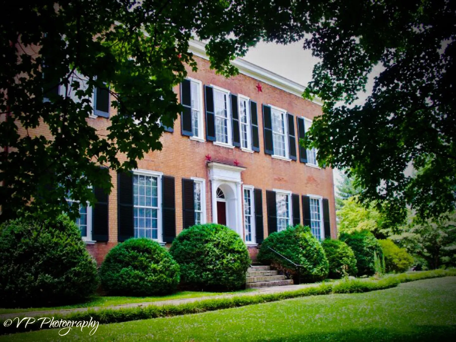 Red brick mansion with black shutters framed by trees and clipped shrubs