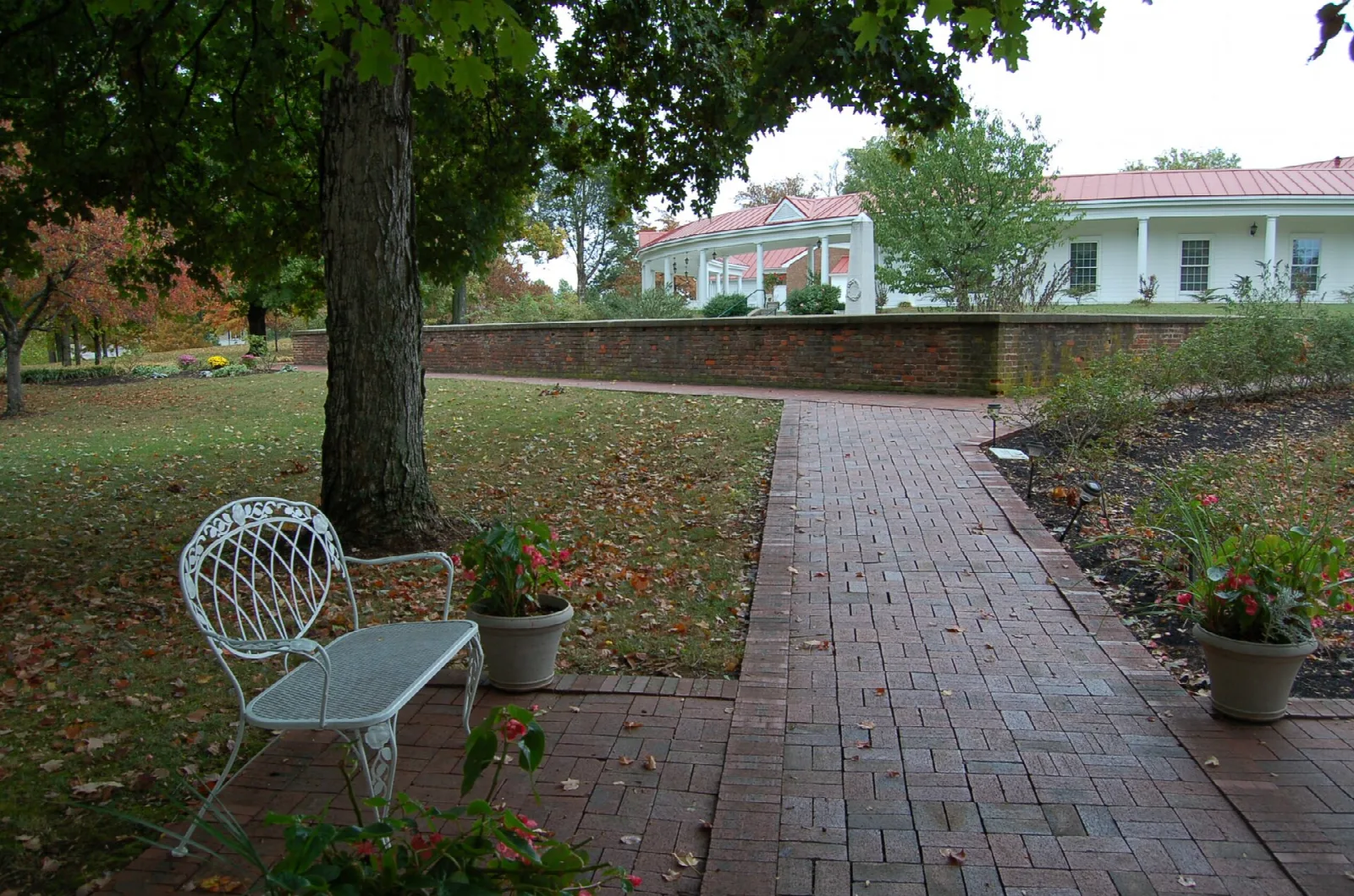 Brick garden path with a white metal bench under a large tree, mansion beyond