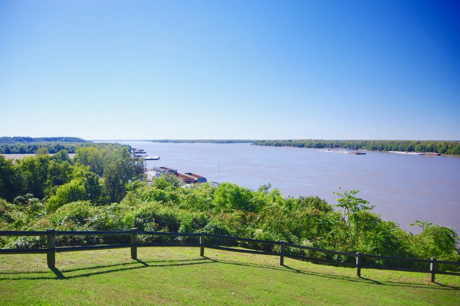Wide brown Mississippi River seen from a grassy bluff with a fence and trees below