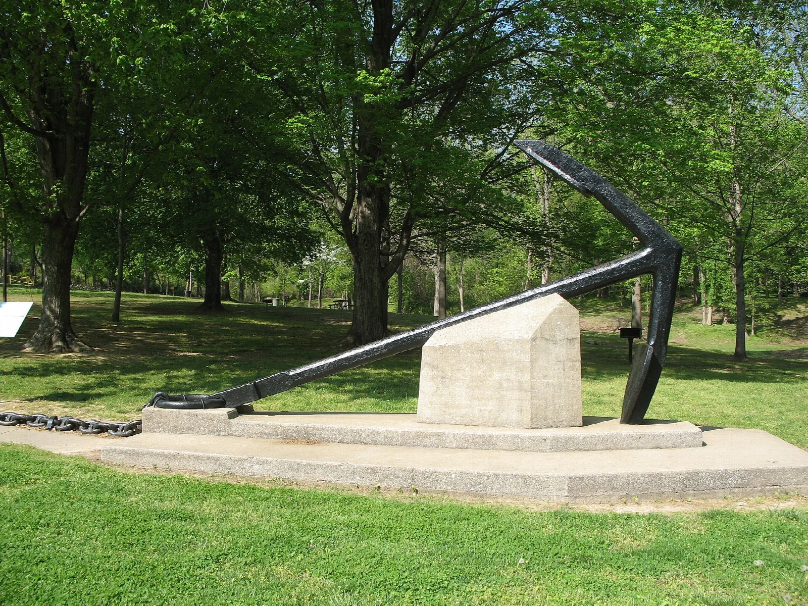 Large black anchor on a stone base on a green lawn under shade trees