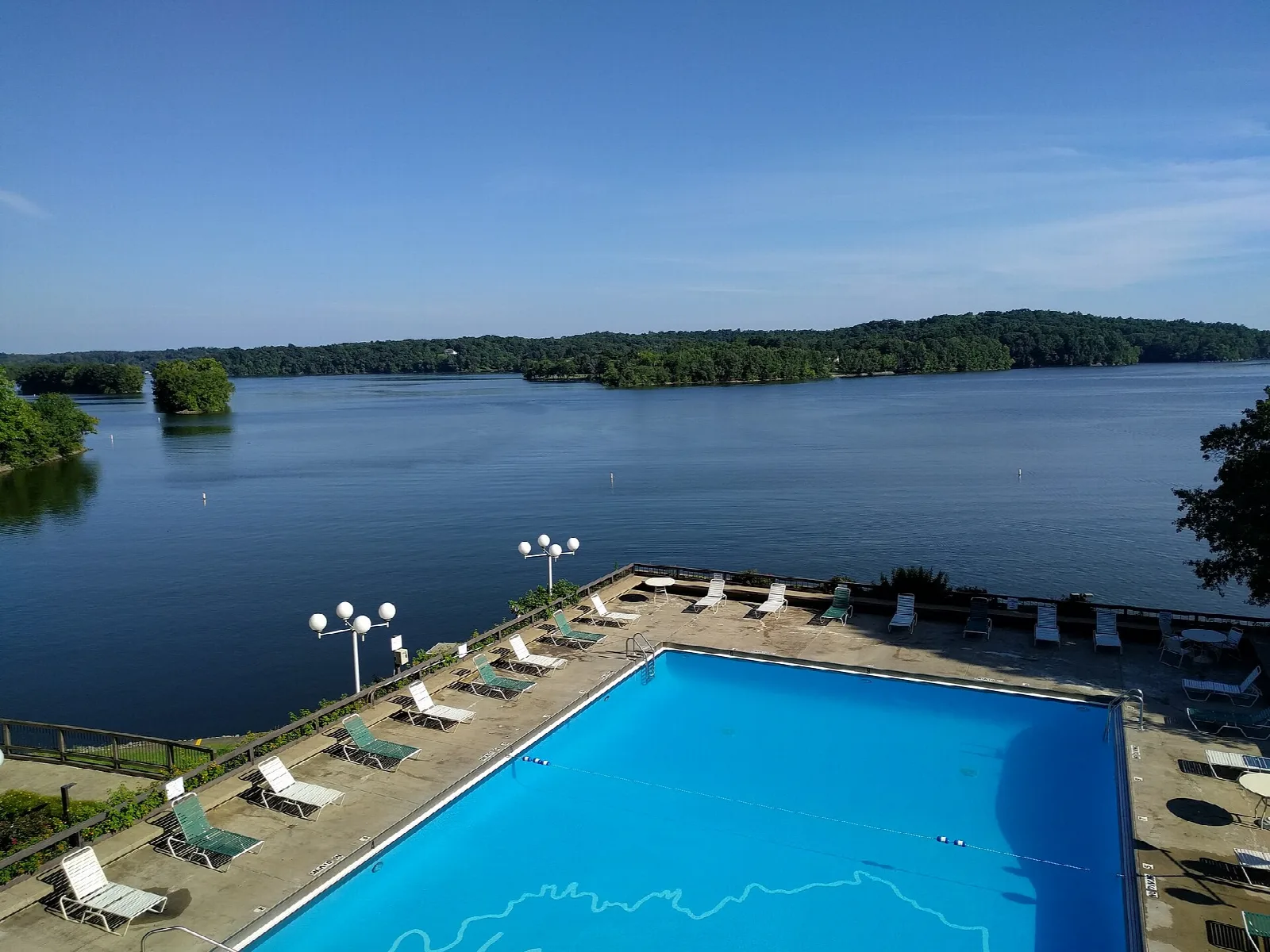 Lake with wooded shore seen from a lodge balcony, with a blue pool and lounge chairs below