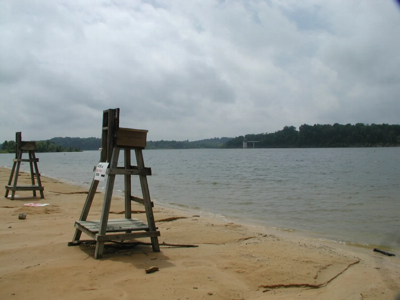 Sandy lakeshore with two wooden lifeguard stands and a wooded far bank under a cloudy sky