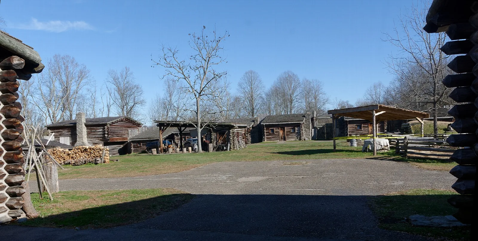 Open fort yard with log cabins and a covered shelter under a blue sky