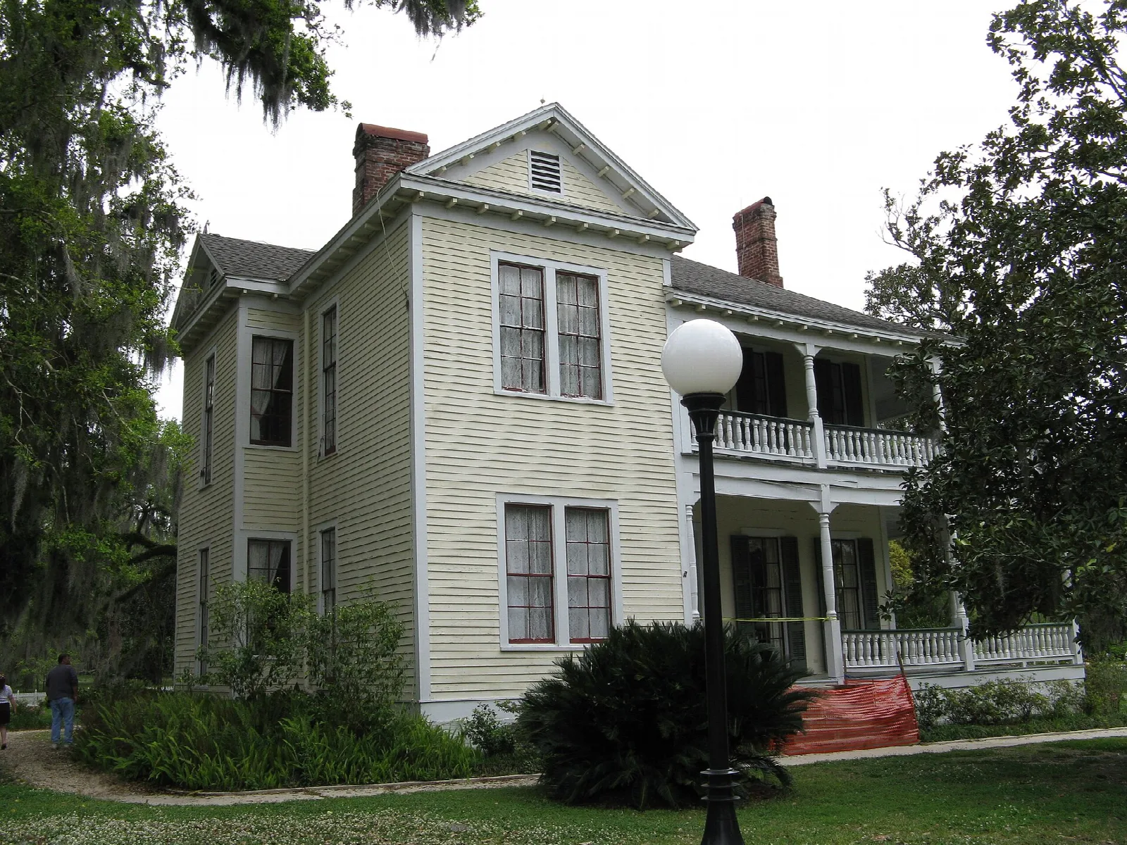 Two-story white house with a covered upper porch and a lamp post, shaded by oaks