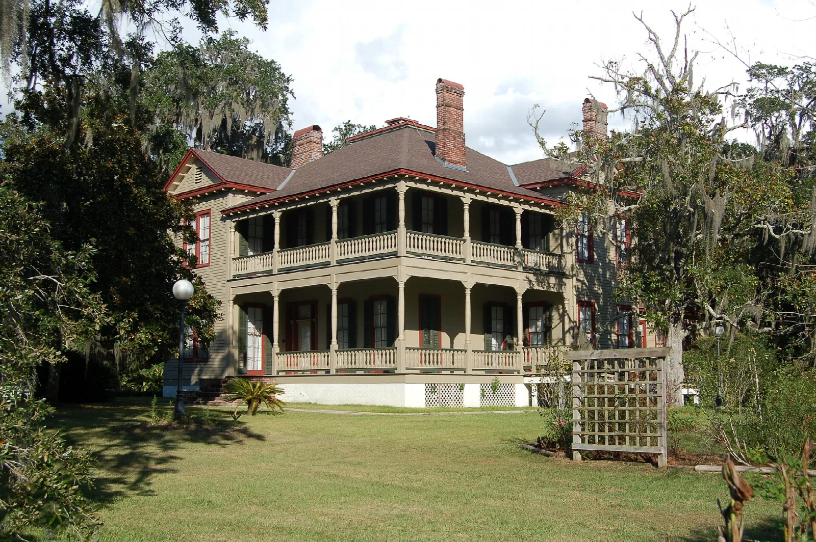 Large house with a wraparound porch behind a green lawn and moss-hung oaks