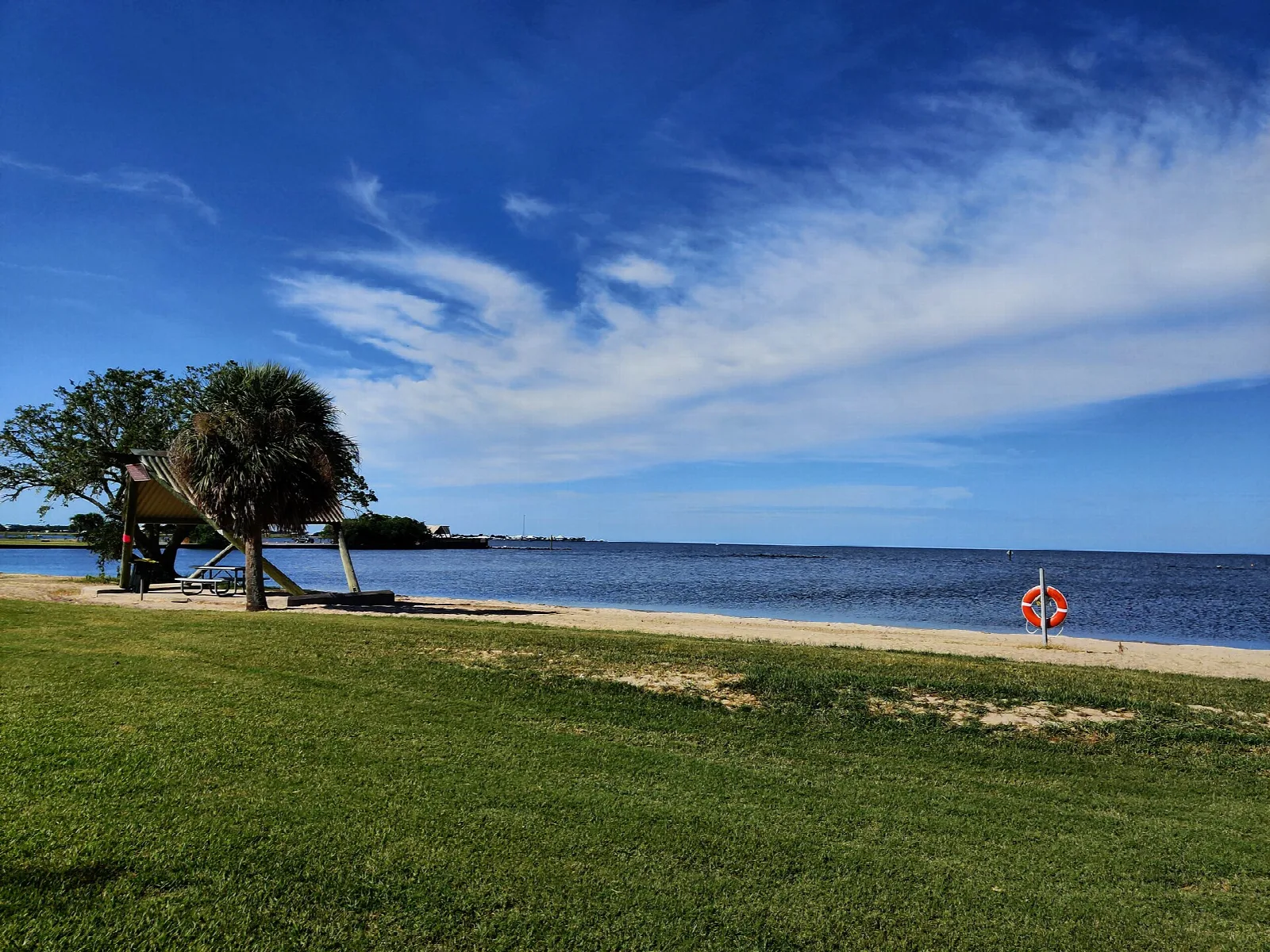 Green lawn and a small beach beside blue bay water, with trees and a life ring on the shore