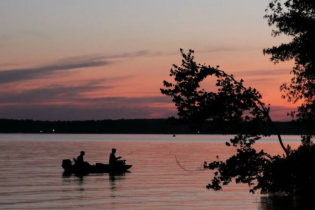 Two anglers in a small boat silhouetted on the lake at sunset, framed by tree branches