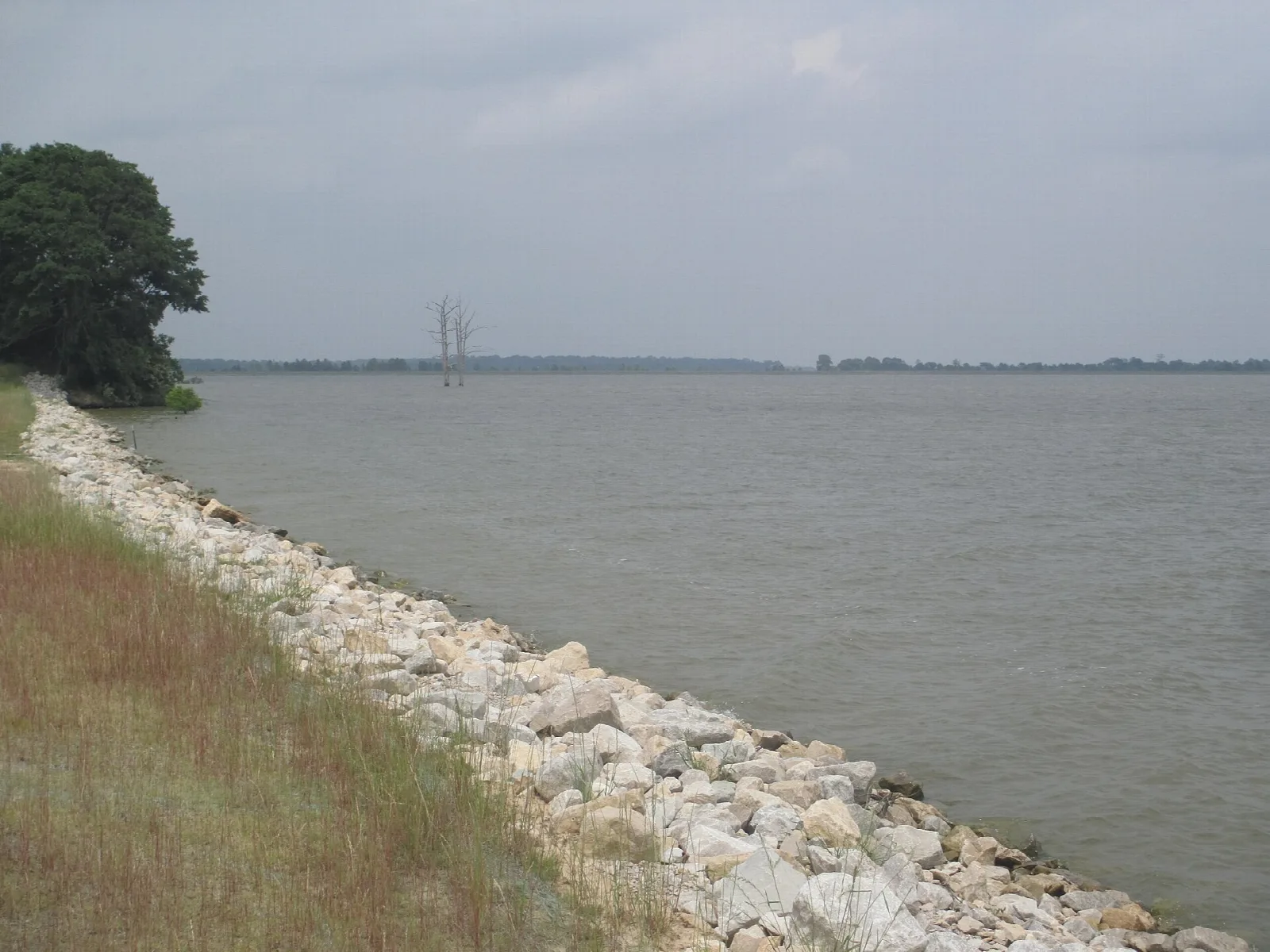 Rock-lined shoreline along a wide gray reservoir under a cloudy sky