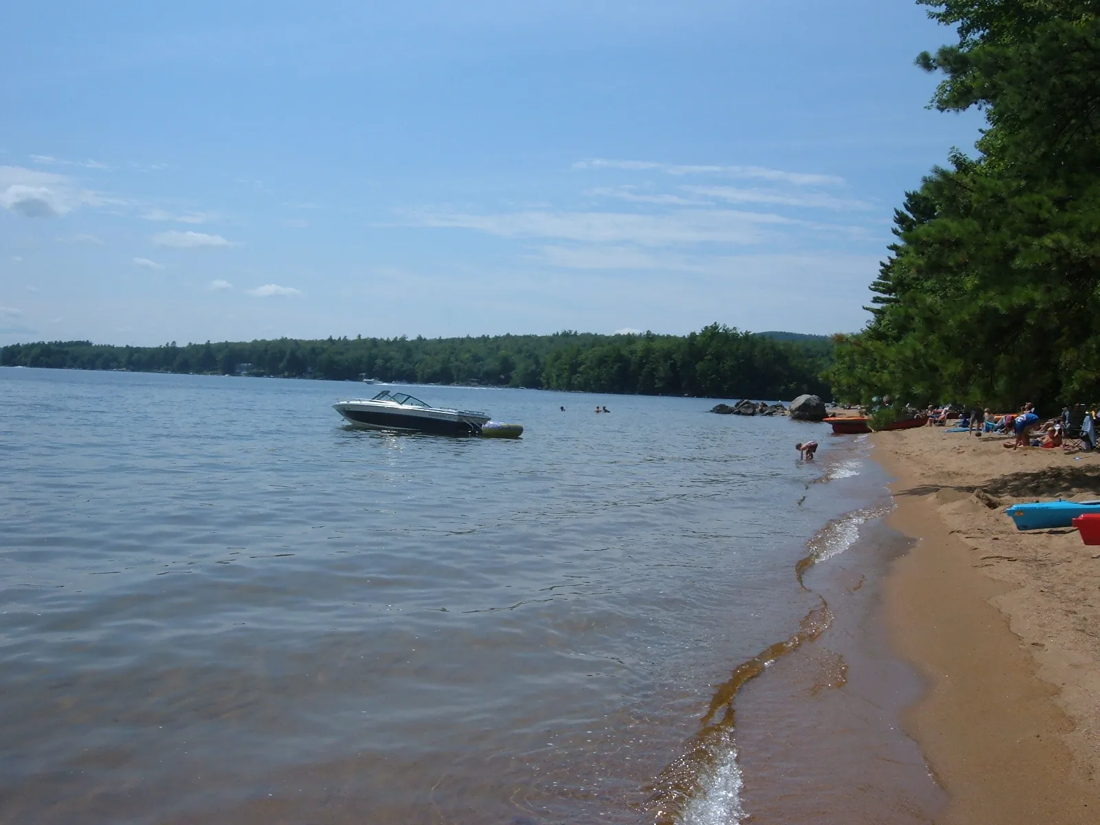 Sandy beach curving along calm lake water with a boat anchored offshore and trees on the point