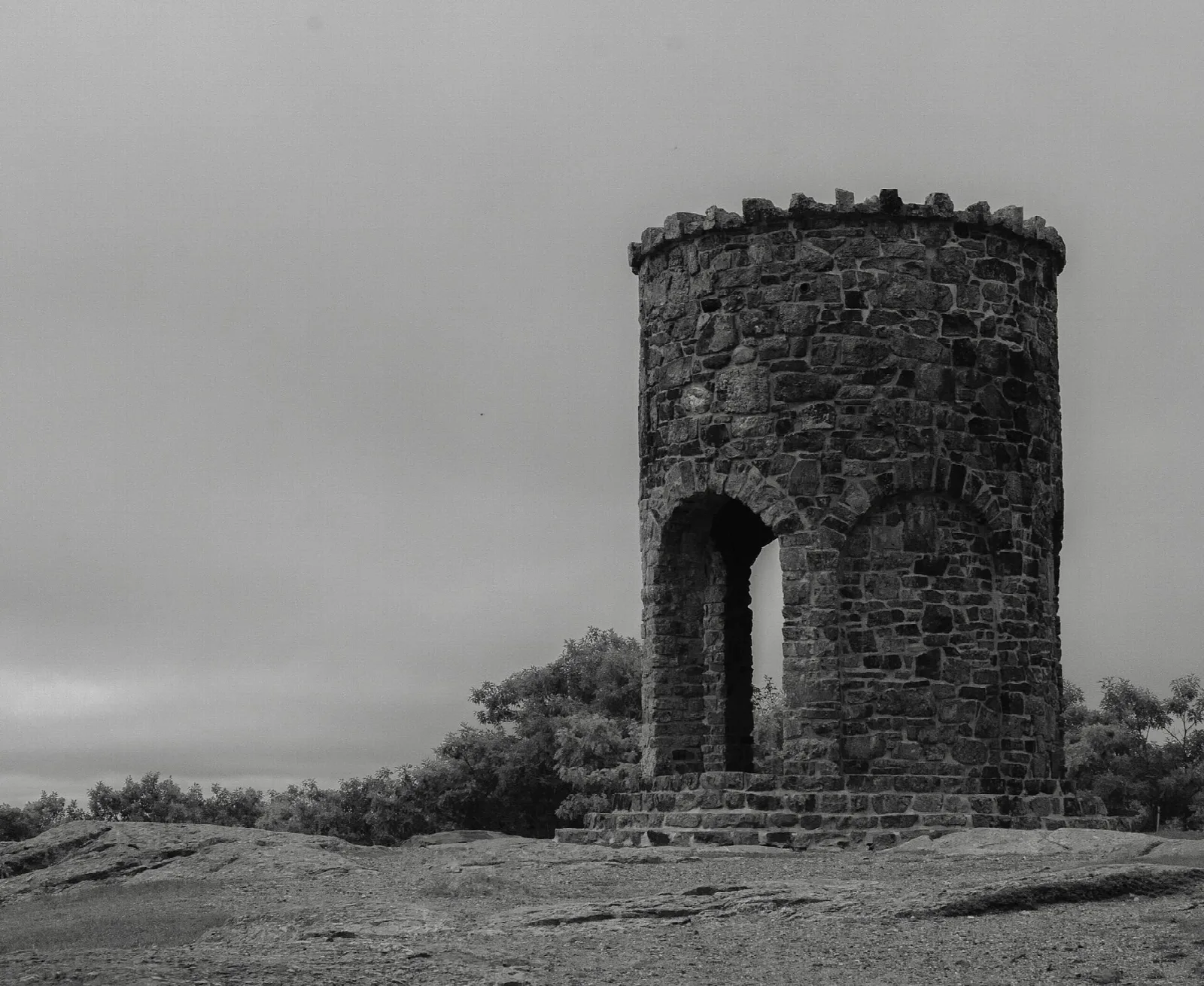 Black and white photo of a round stone tower with an arched doorway on a rocky summit