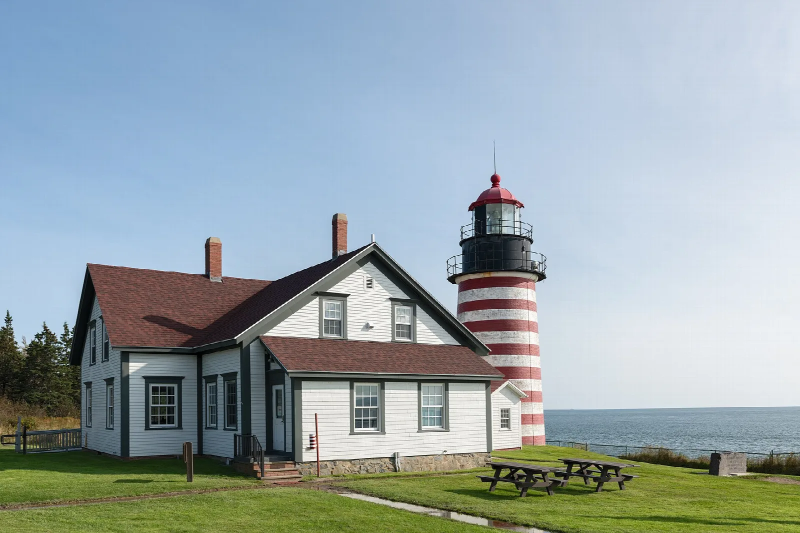 Red-and-white striped lighthouse beside a white house on a green lawn with picnic tables
