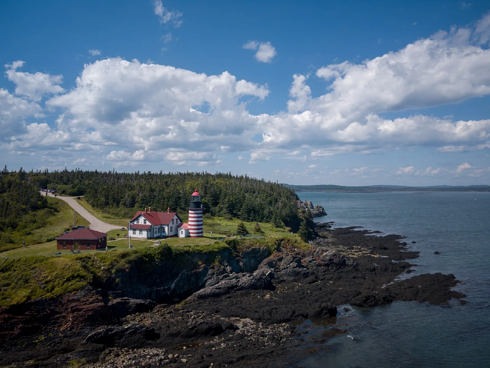 Aerial view of a striped lighthouse and keeper's house on a green bluff above dark rocky shore