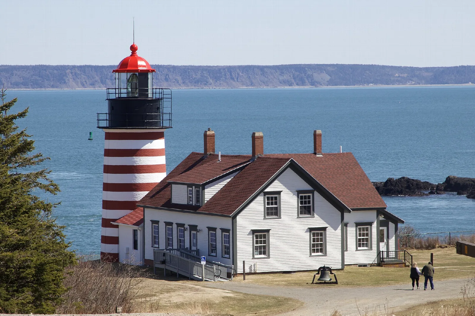 Striped lighthouse and white house beside blue water with distant cliffs across the channel