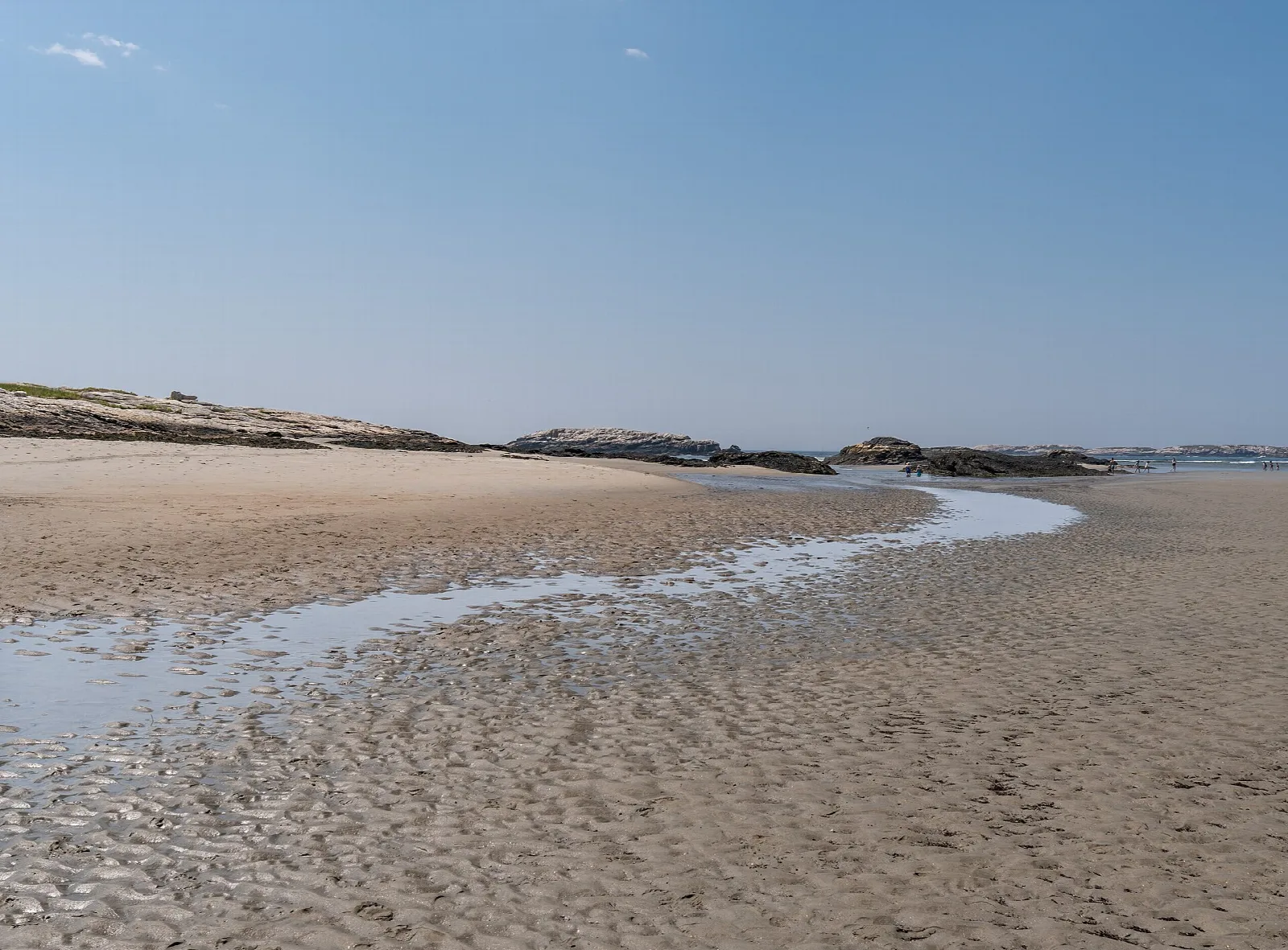 Wide tidal sand flat with a shallow channel and rocks in the distance