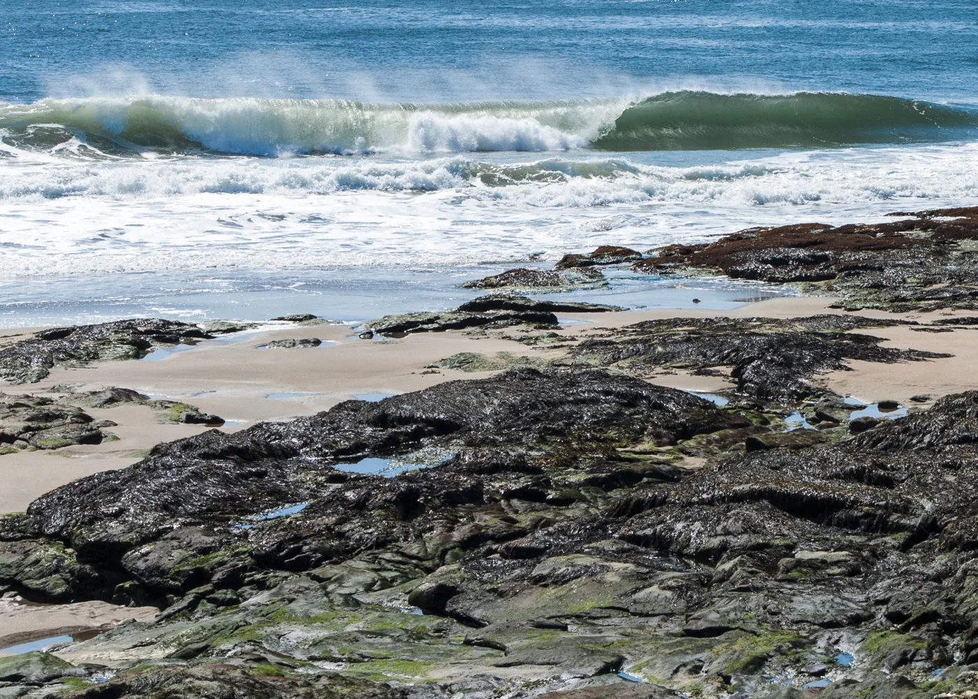 Waves breaking over dark wet rocks and sand under a blue sky